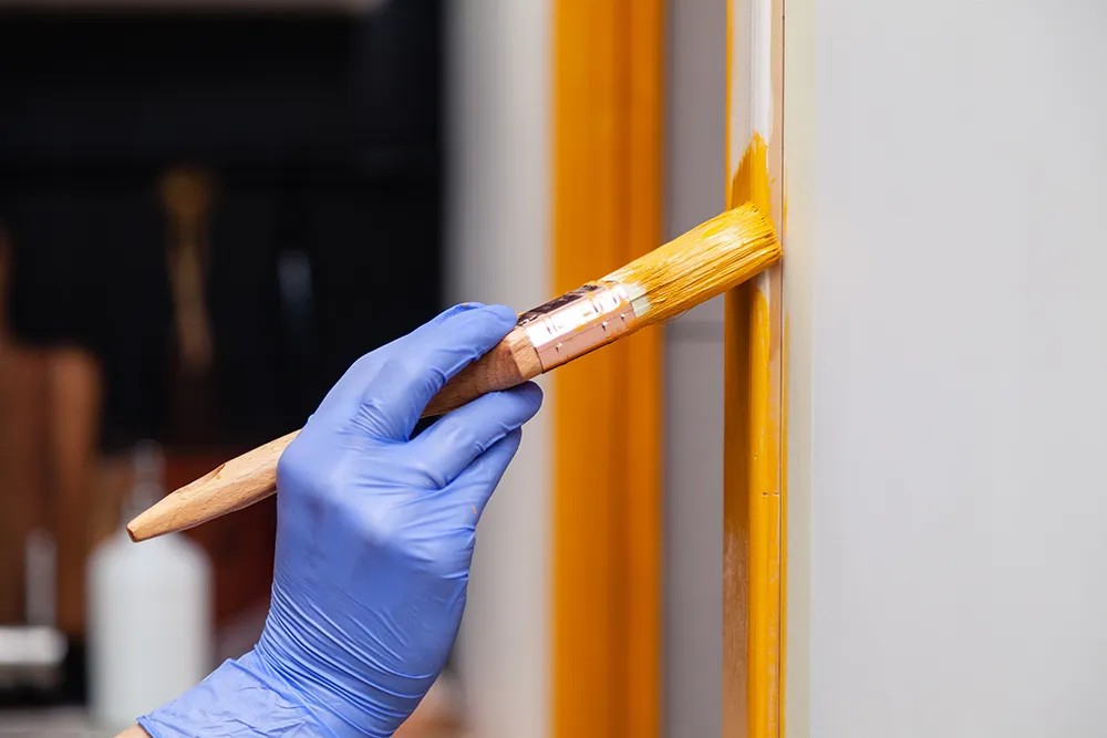 Gros plan sur une main de femme portant un gant en caoutchouc violet et tenant un pinceau, en train de peindre une porte en bois naturel avec de la peinture orange ; design créatif et lumineux pour un intérieur ; comment peindre une surface en bois